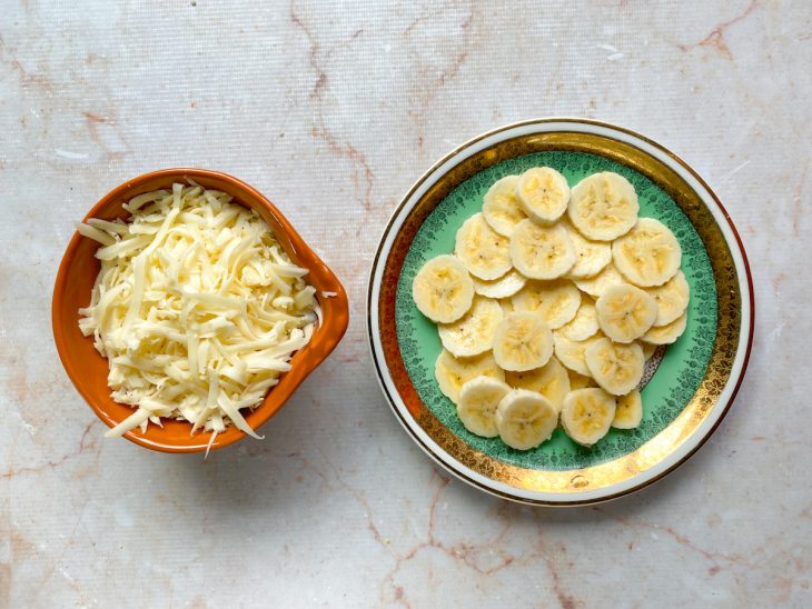 Queijo ralado e banana em rodelas reservados em recipientes distintos.
