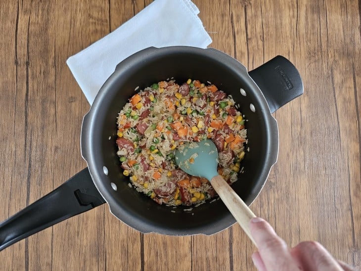 Uma panela de pressão com calabresa, cebola, pimentão, cenoura, milho, ervilha e arroz.