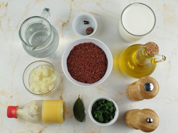 Bancada organizada com o arroz vermelho, leite, temperos e a cebola ralada em recipientes separados.