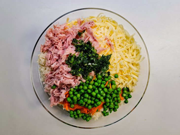 Arroz cozido, cenoura, cebola, queijo mussarela, presunto, ervilha, cheiro-verde e cebolinha em um refratário de vidro.