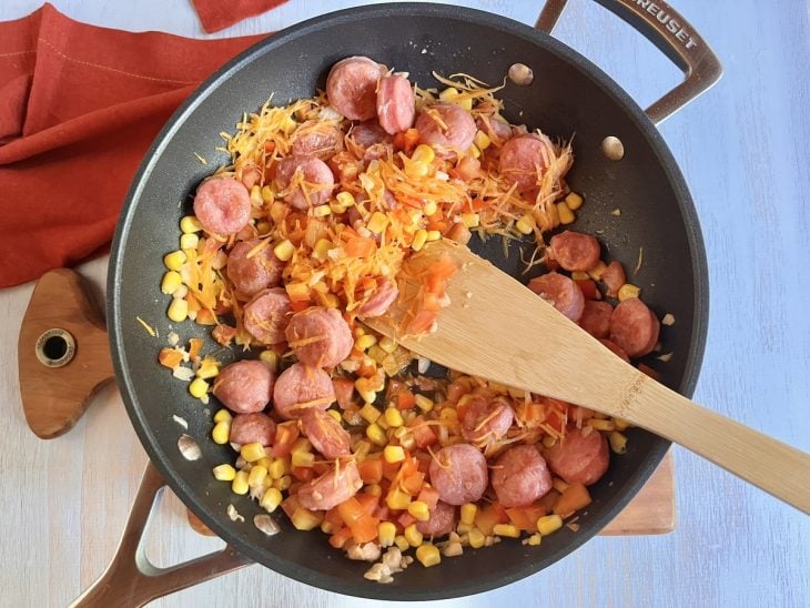 Uma panela refogando rodelas de linguiça e cebola, pimentão, tomate, milho e cenoura.