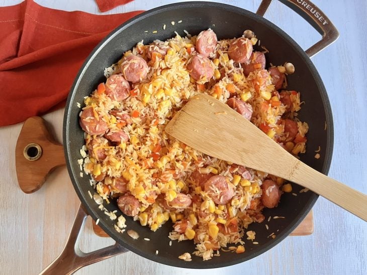 Uma panela refogando rodelas de linguiça e cebola, pimentão, tomate, milho, cenoura e arroz.