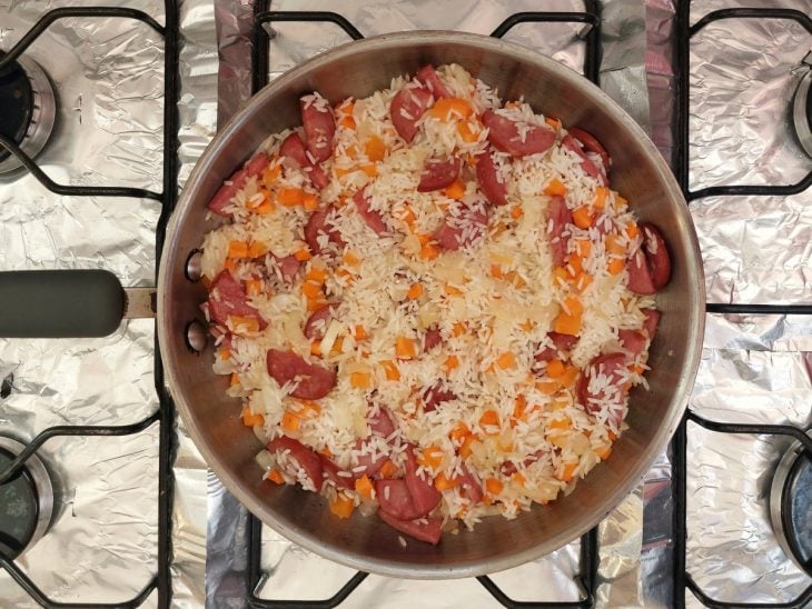 Uma panela refogando calabresa, cebolas, legumes e arroz.