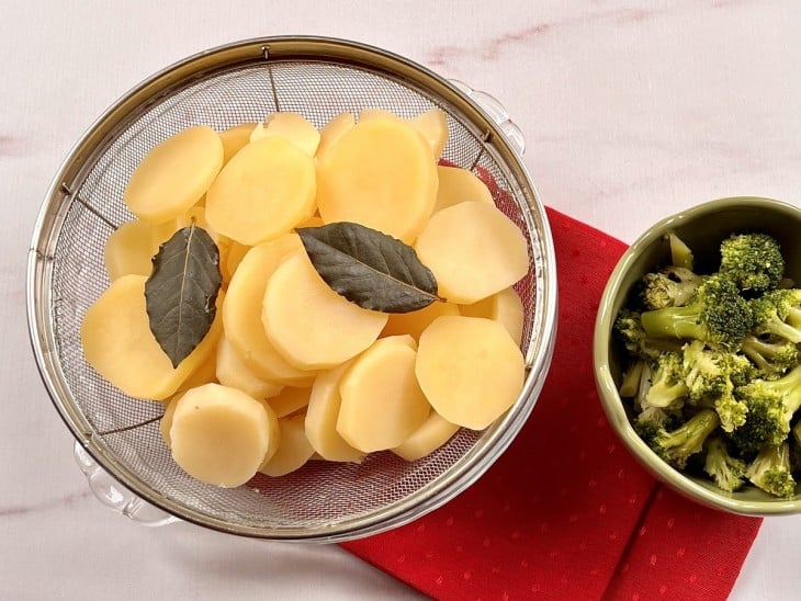 Dois recipientes contendo rodelas de batata cozidas e outro com brócolis cozidos.