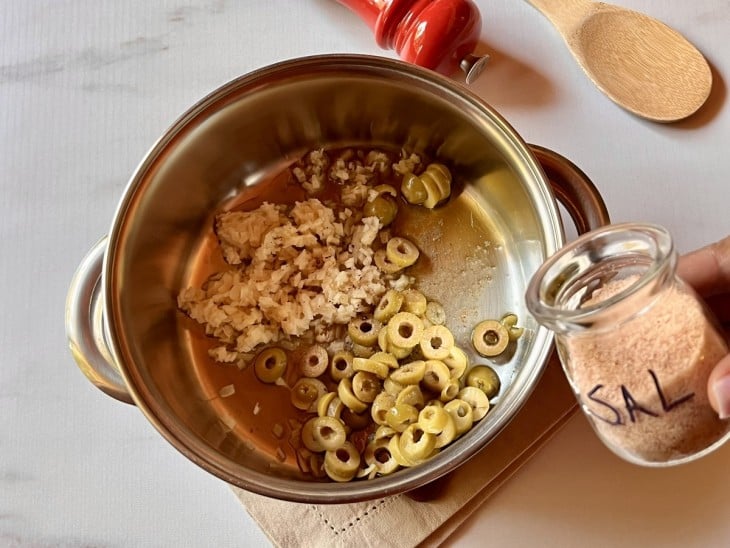 Uma panela refogando, no azeite, cebolas e azeitonas com pimenta e sal.