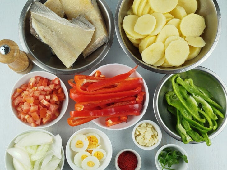 Ingredientes organizados na bancada com batatas, tomates, pimentões, cebolas, alho, ovos e bacalhau preparados para o uso.
