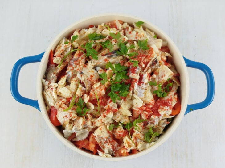 Pedaços de bacalhau, salsa, alho laminado e colorau sendo distribuídos sobre as camadas.