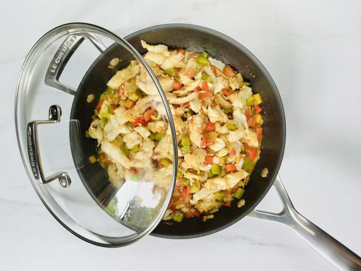 Uma frigideira com alho, cebola, pimentões, tomate, bacalhau e temperos cozinhando na água.