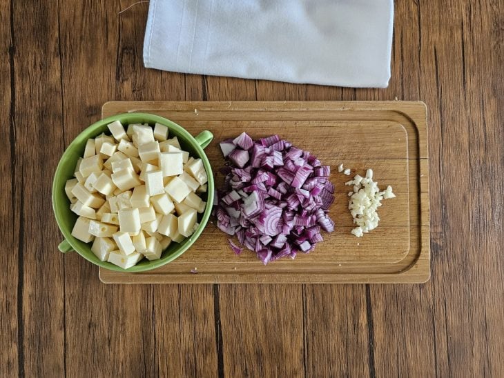 Uma tábua contendo queijo coalho em cubos, cebola-roca picada e alho picado.