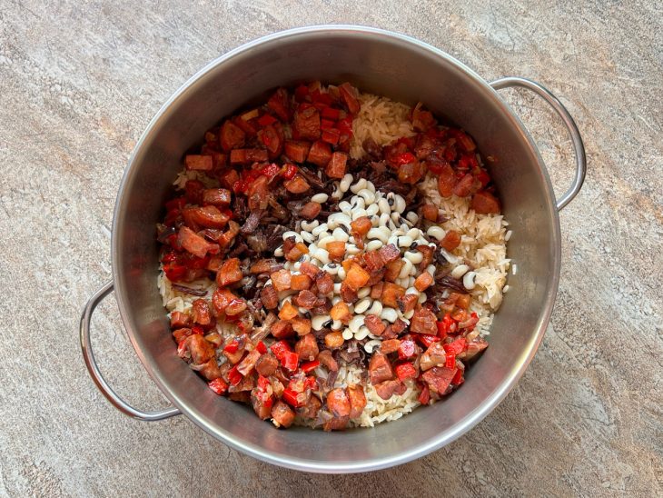 Na panela do arroz, estão acrescentados o feijão, a carne seca, o bacon, a calabresa, o pimentão e a cebola.