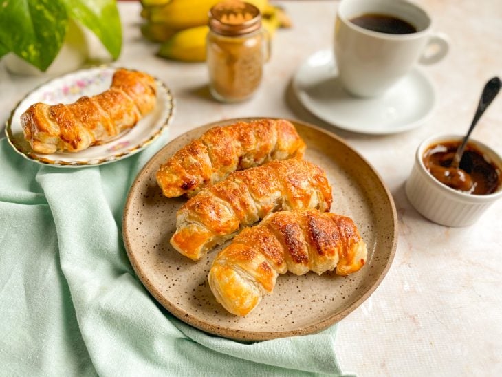 Bananas folhadas prontas para consumo, acompanhadas por café e doce de leite.