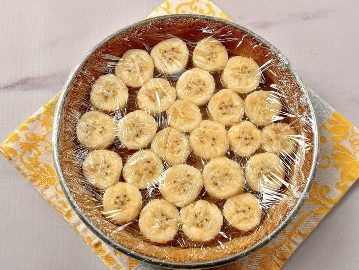 Uma forma contendo massa com doce de leite e rodelas de banana, coberto com plástico filme.