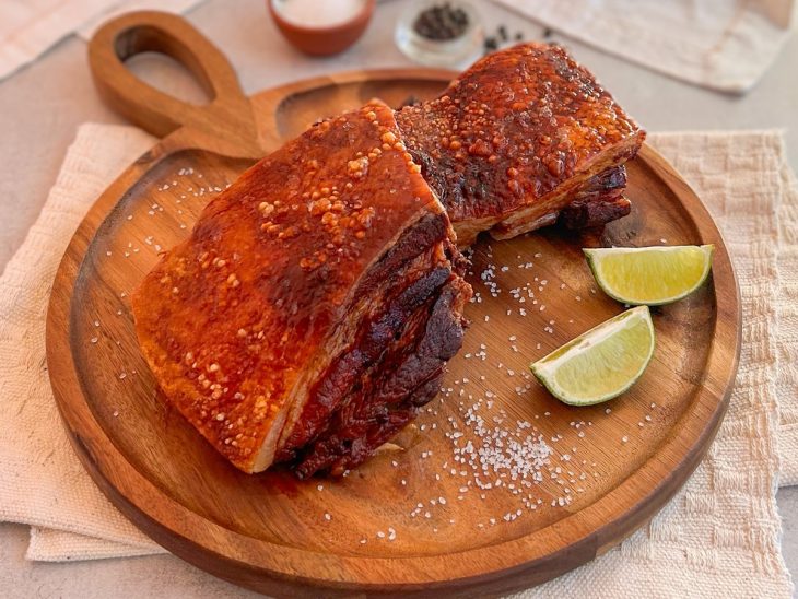 Peça da barriga de porco pronta, posicionada em uma tábua de servir com limão acompanhando.