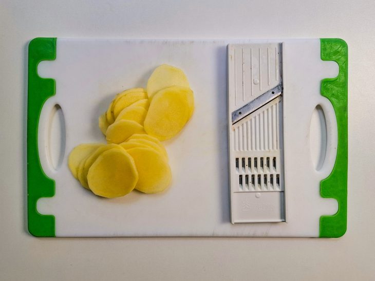 Batatas fatiadas sobre uma tábua ao lado de um mandolim.
