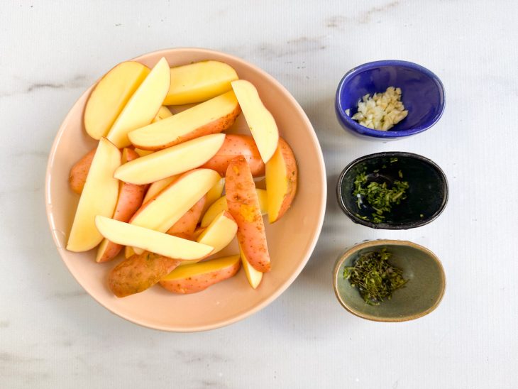 Batatas cortadas em gomos grandes com ervas e alho picados ao lado.