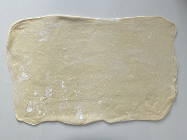 Rolo de madeira abrindo a massa crescida na bancada em formato de um retângulo grande.