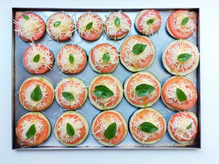 Uma assadeira com rodelas de tomate sobre rodelas de berinjelas salpicadas com parmesão e manjericão.