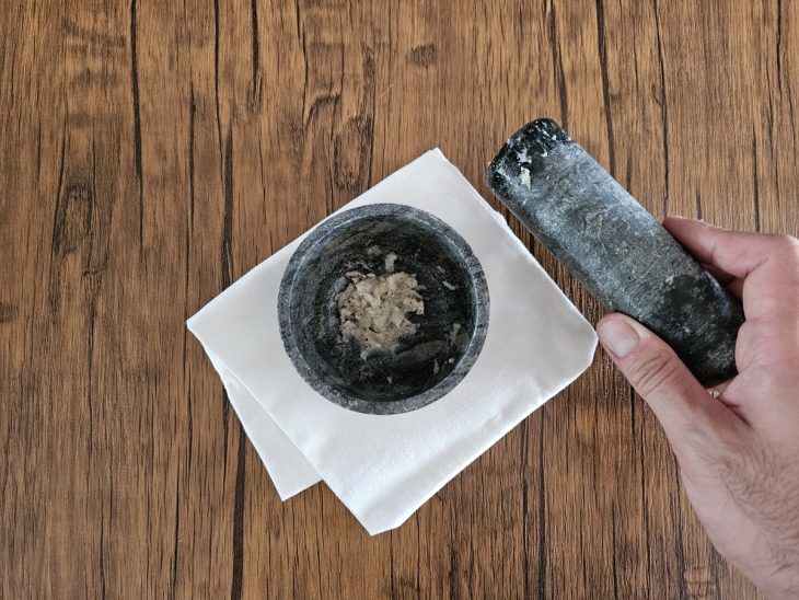 Um pilão contendo uma pastinha de sal, alho descascado e pimenta-do-reino em grãos.