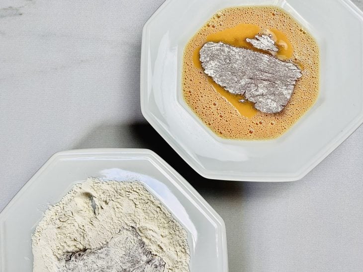 Um prato branco com um bife sobre os ovos batidos e outro com um bife sobre a farinha de trigo.