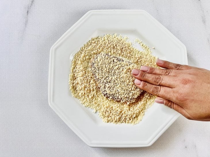 Bife em um prato com farinha de rosca e manuseado com uma mão.