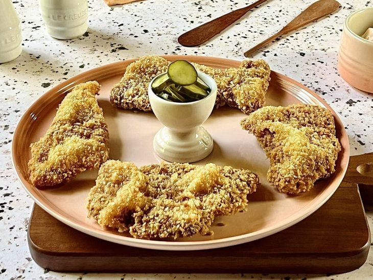 Bifes à milanesa crocantes prontos servidos em um prato redondo rosa, acompanhados por rodelas de pepinos.