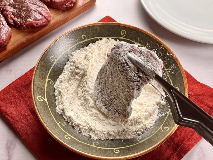 Um bife sendo empanado na farinha.