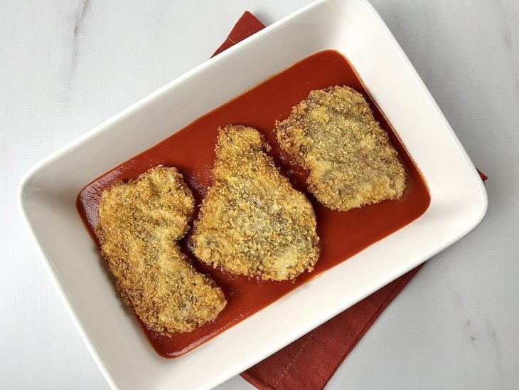 Um refratário forrado com molho de tomate e contendo 3 bifes empanados.