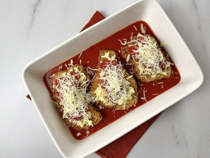 Um refratário forrado com molho de tomate e contendo 3 bifes empanados cobertos com queijo ralado.