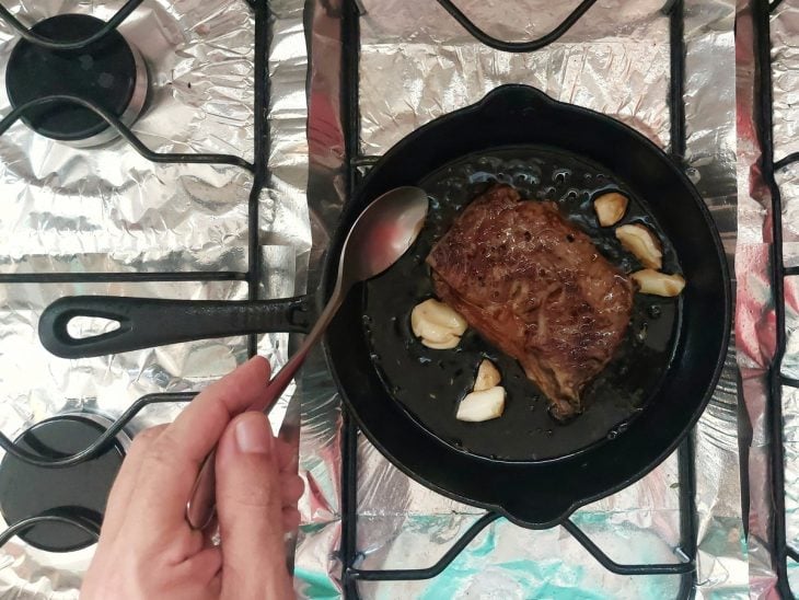 Bife na frigideira sendo regado com a gordura aromatizada com alho.