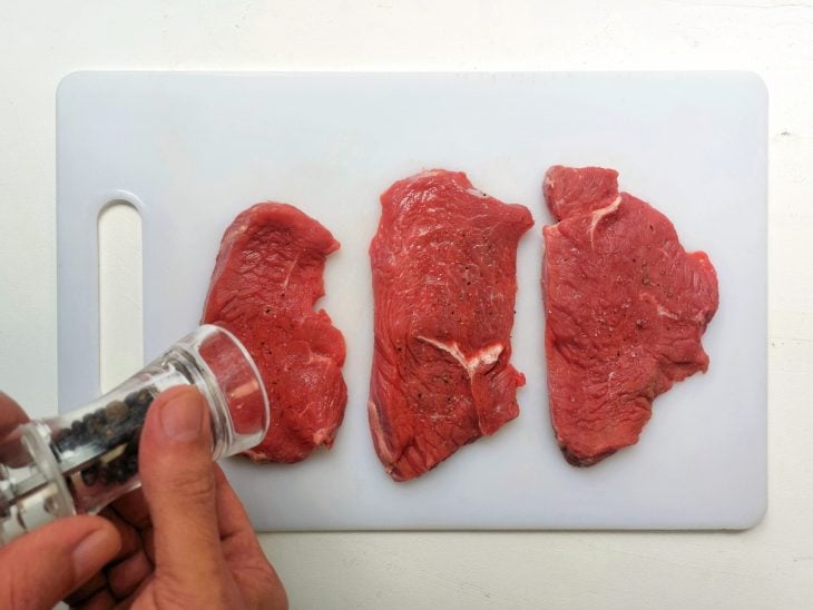 Mão com moedor de pimenta temperando os bifes na tábua.