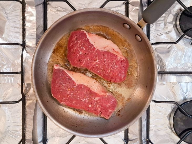 Sal e pimenta-do-reino adicionados sobre os bifes na frigideira.