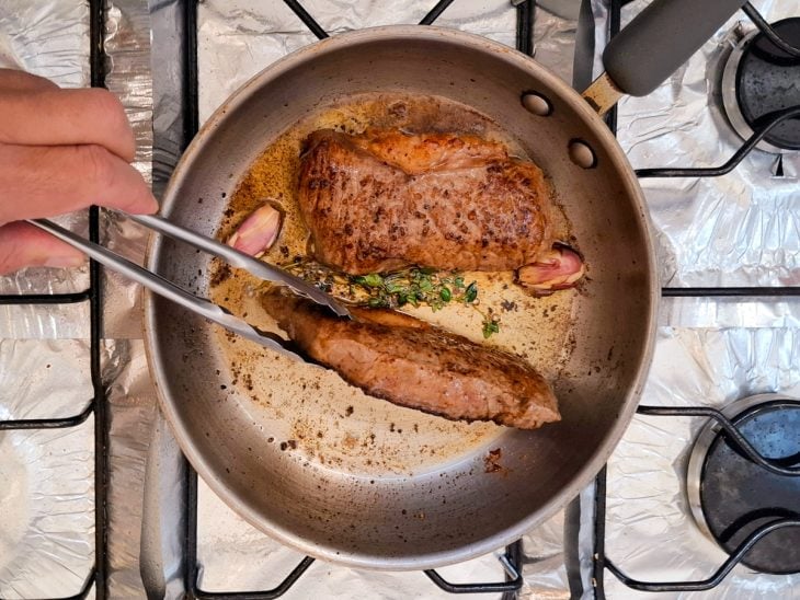 Bife posicionado verticalmente com um pegador para fritar a gordura na frigideira.