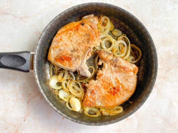 Uma frigideira com as bistecas e as rodelas de cebola caramelizadas.