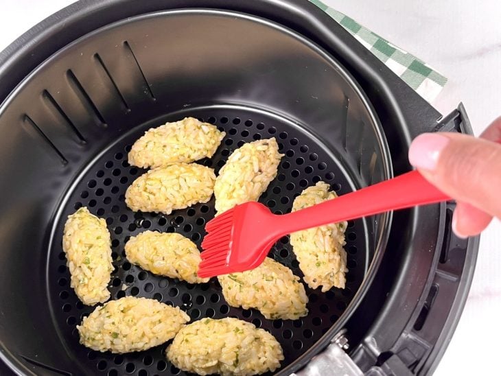 Uma cesta de airfryer contendo bolinhos de arroz crus sendo pincelados com azeite.