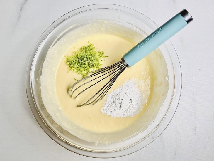 Mistura do liquidificador misturado com farinha, fermento e raspas de limão em uma tigela de vidro.