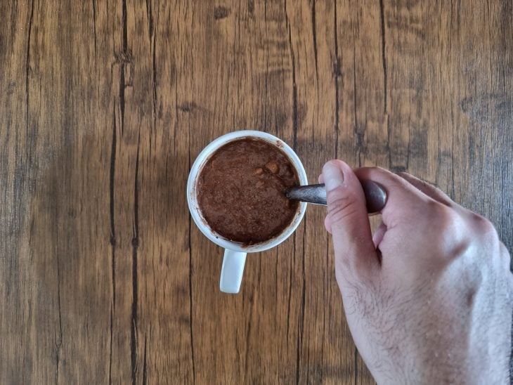 Caneca branca com massa do bolo sendo misturada com o auxílio de uma colher.