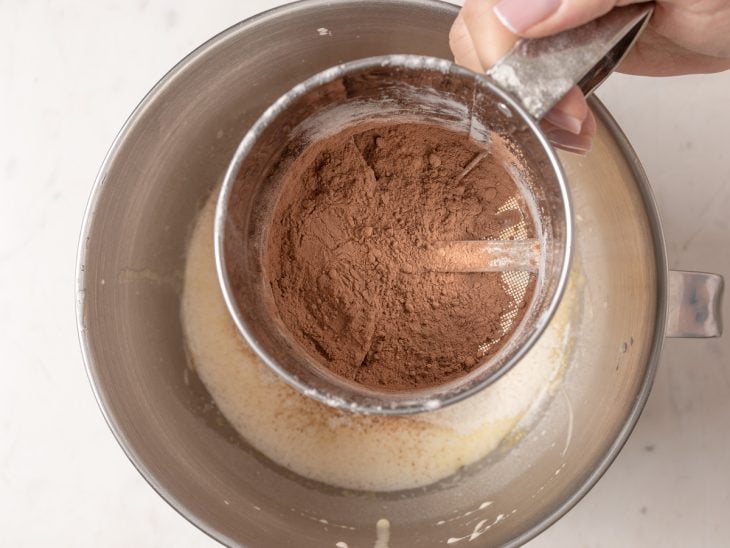 Uma tigela com ovos, açúcar, óleo, leite, farinha e chocolate em pó peneirado.