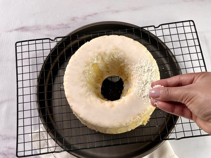 Uma forma contendo bolo de coco com cobertura e sendo decorando com coco ralado.