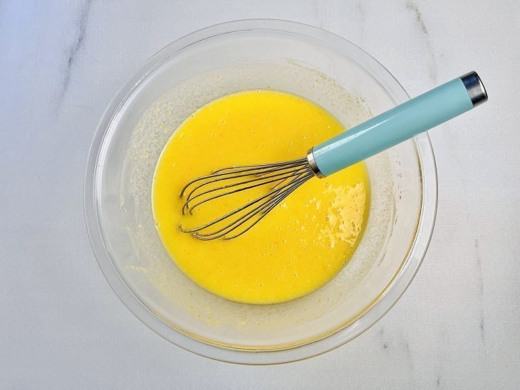 Ingredientes batidos em uma tigela de vidro com auxílio de um fouet.