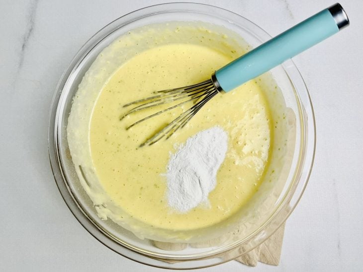 Mistura do liquidificador, farinha e fermento adicionados em um bowl de vidro.