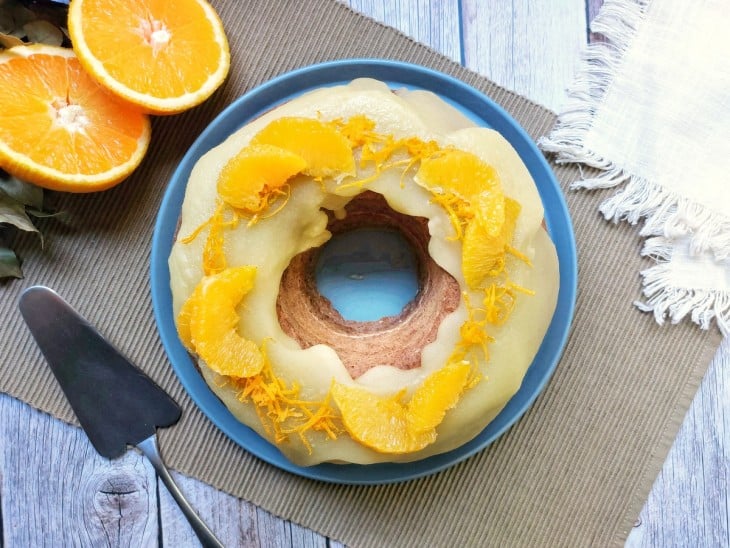 Bolo pronto com a cobertura durinha pro cima. Disposto em um prato de cor azul grande e com pedaços de laranja para decorar.