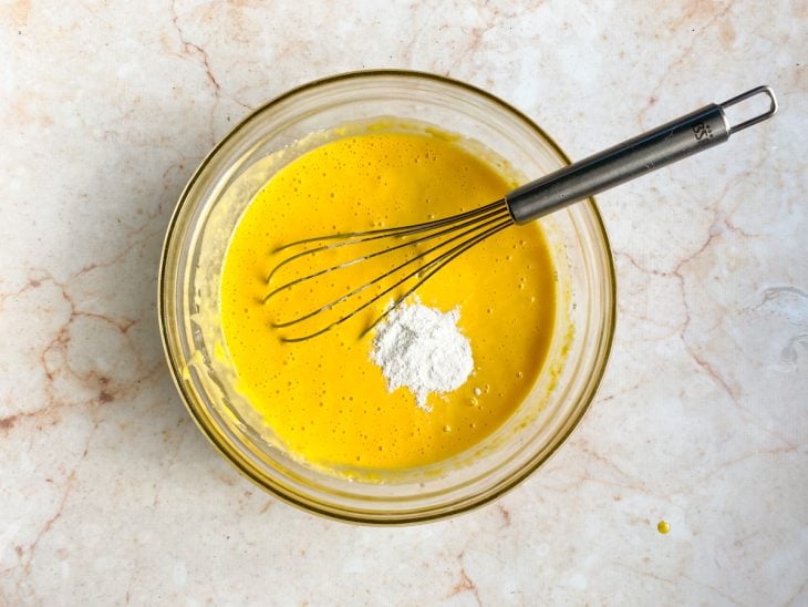 Uma tigela com ovos, açúcar, suco de laranja, óleo, farinha e fermento batidos com fouet.