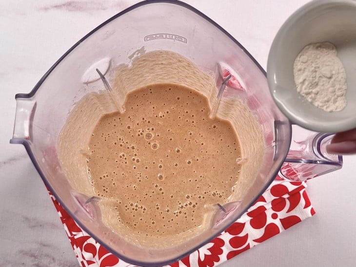 Um liquidificador com a mistura de ovos, maçãs, açúcar, canela em pó, noz-moscada, óleo, sal, amido de milho, quinoa, farinha de arroz e goma xantana.