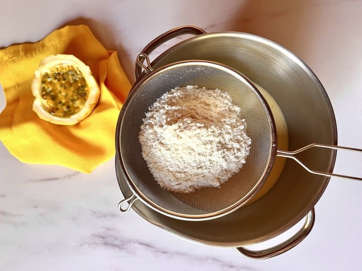 Tigela com a mistura dos ingredientes líquidos da massa, com uma peneira por cima peneirando a farinha.