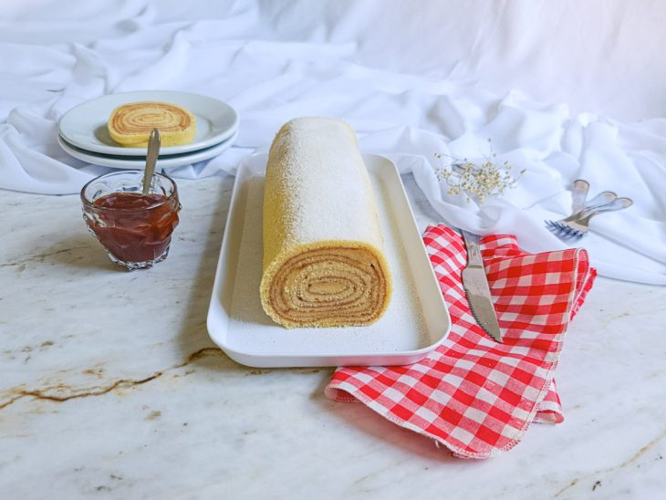 Bolo de rolo pronto para consumo com fatia cortada e servida num prato de sobremesa ao fundo.