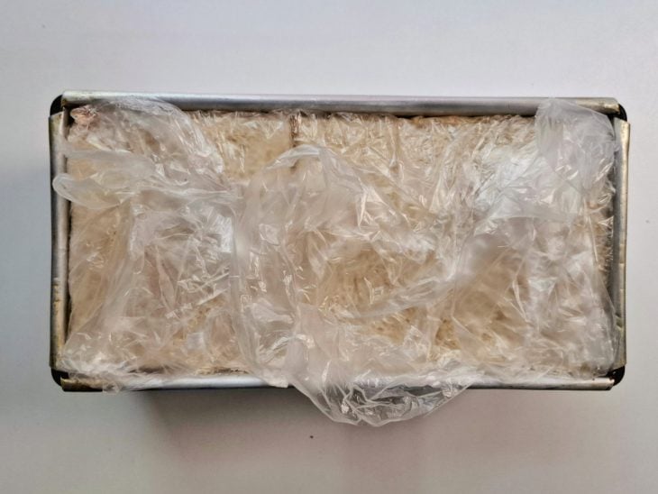 Camadas de recheio e de pão adicionados a forma e plástico cobrindo a superfície.