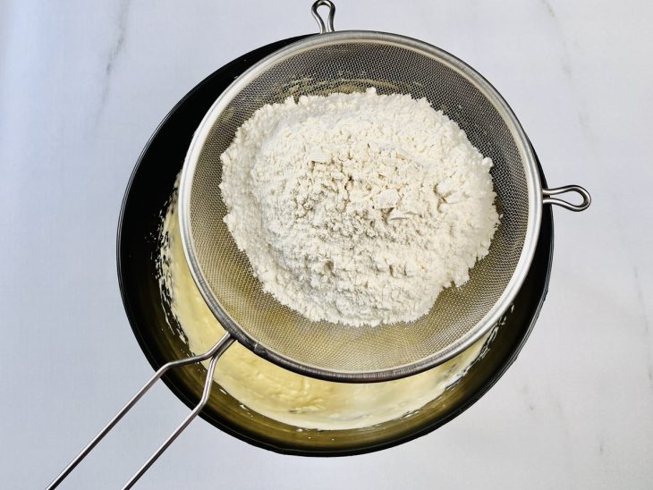 Na massa batida, farinha de trigo sendo peneirada por cima.