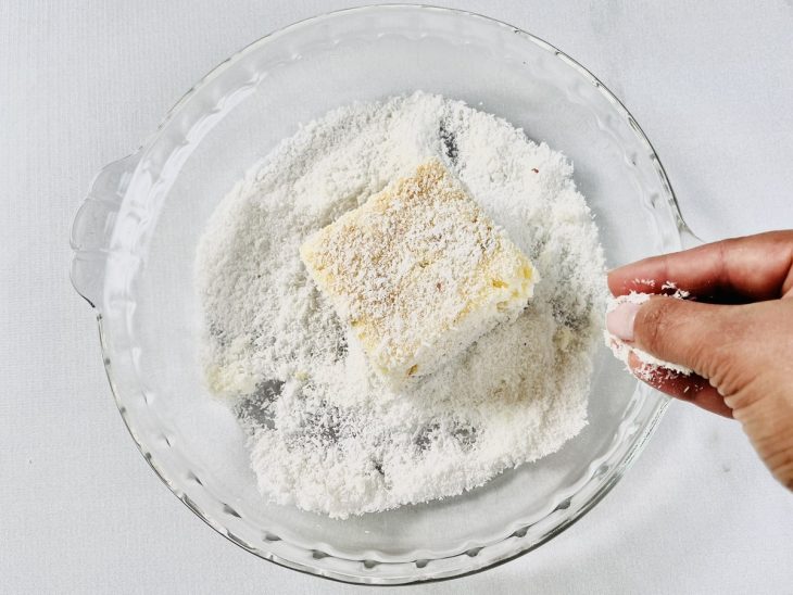 Pedaço do bolo molhado na calda, sendo empanado no coco ralado.