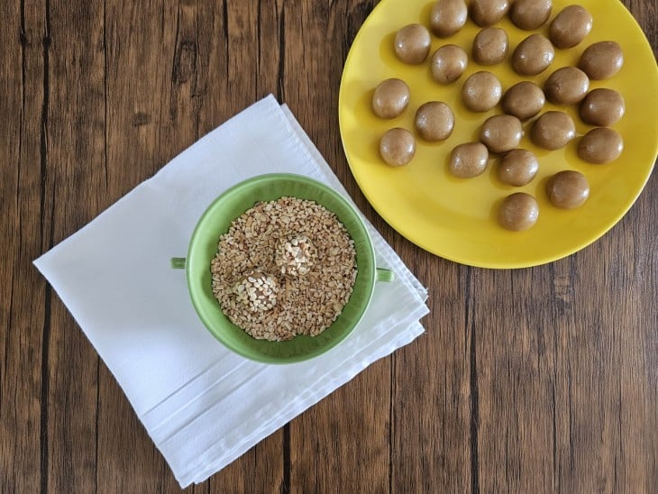 Dois brigadeiros de amendoim sendo empanados com amendoins triturados.