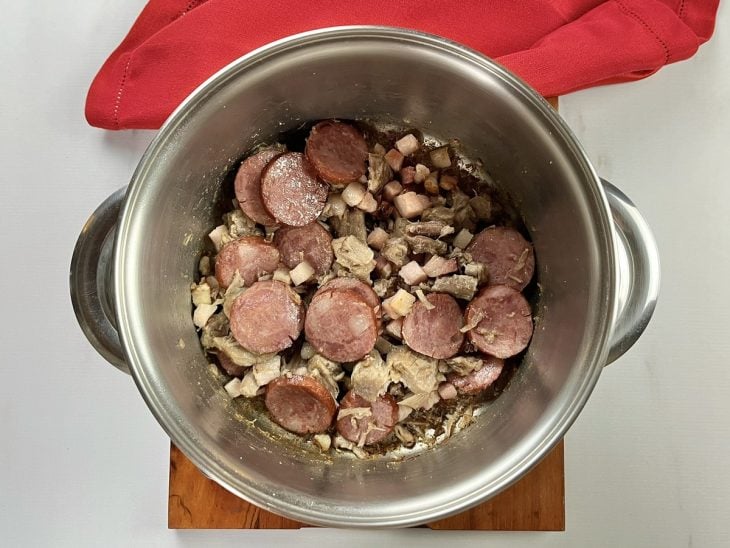 Uma panela com azeite, paio, costelinhas, bacon e cebolas.